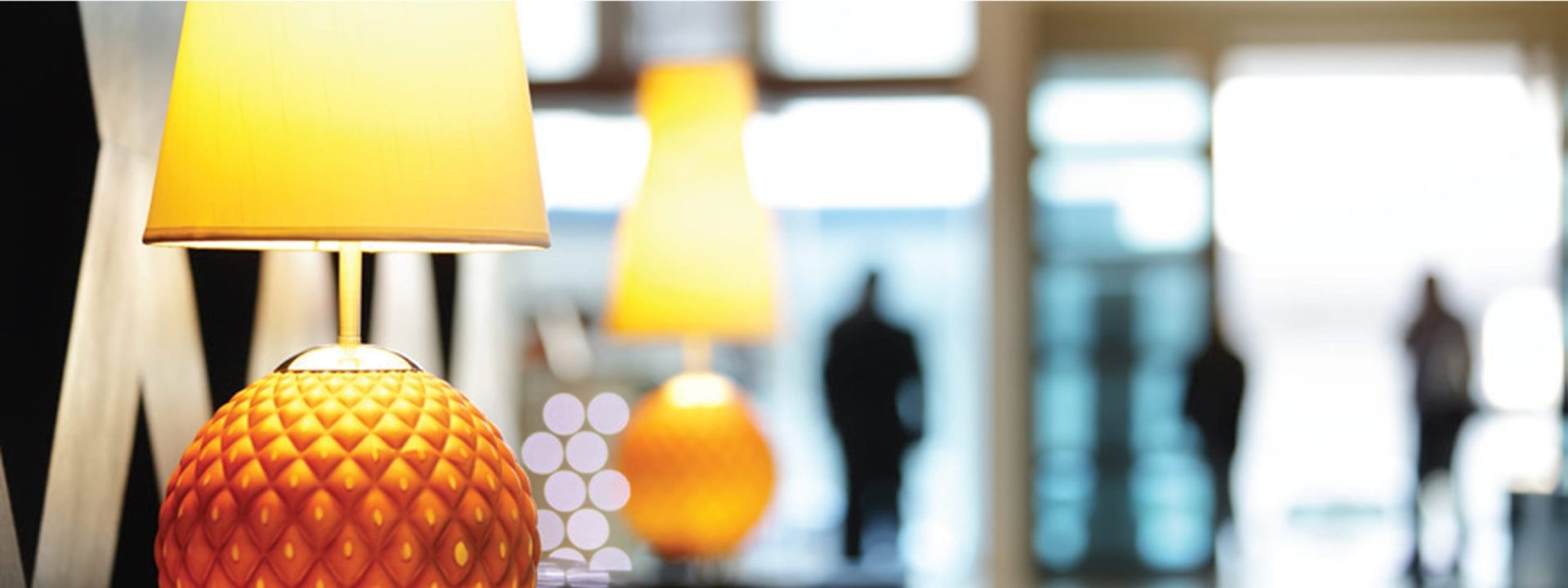 Warmly lit hotel lobby with designer table lamps and guests arriving in the background.