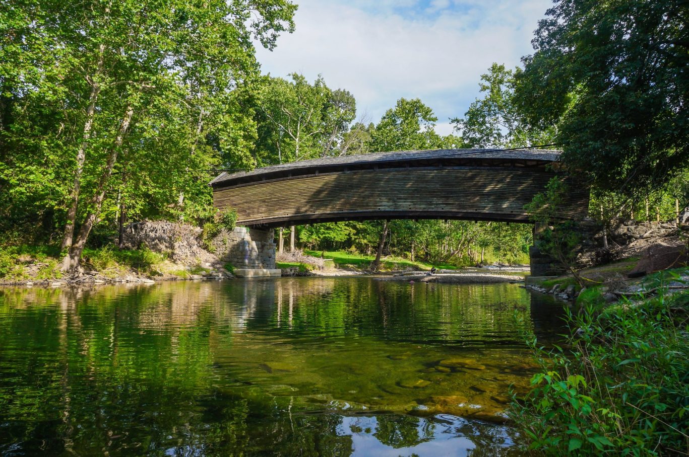 Rustic covered bridge crossing reflective river surrounded by green forest.