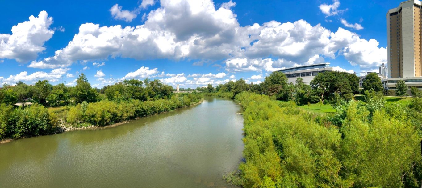 Scenic river view with lush greenery and city buildings under a blue sky in Mansfield, Ohio.