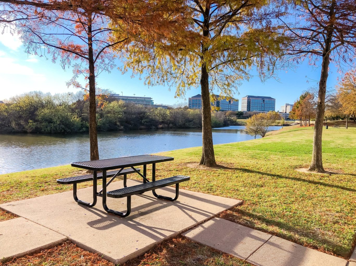 Shaded riverside picnic area in Las Colinas urban district with office buildings in background.