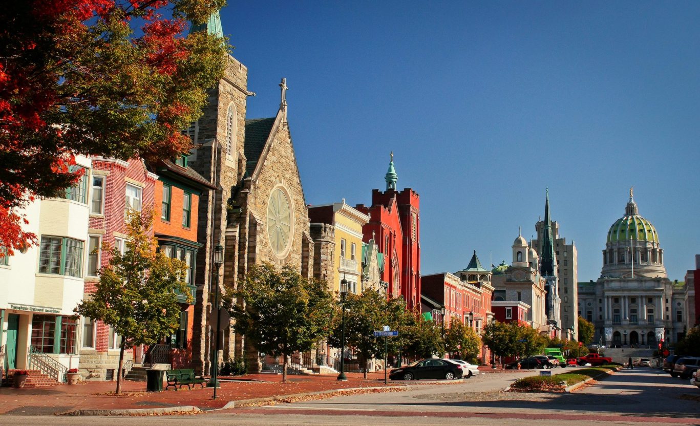 Colorful downtown Harrisburg street with historic buildings and Pennsylvania State Capitol dome in the background.