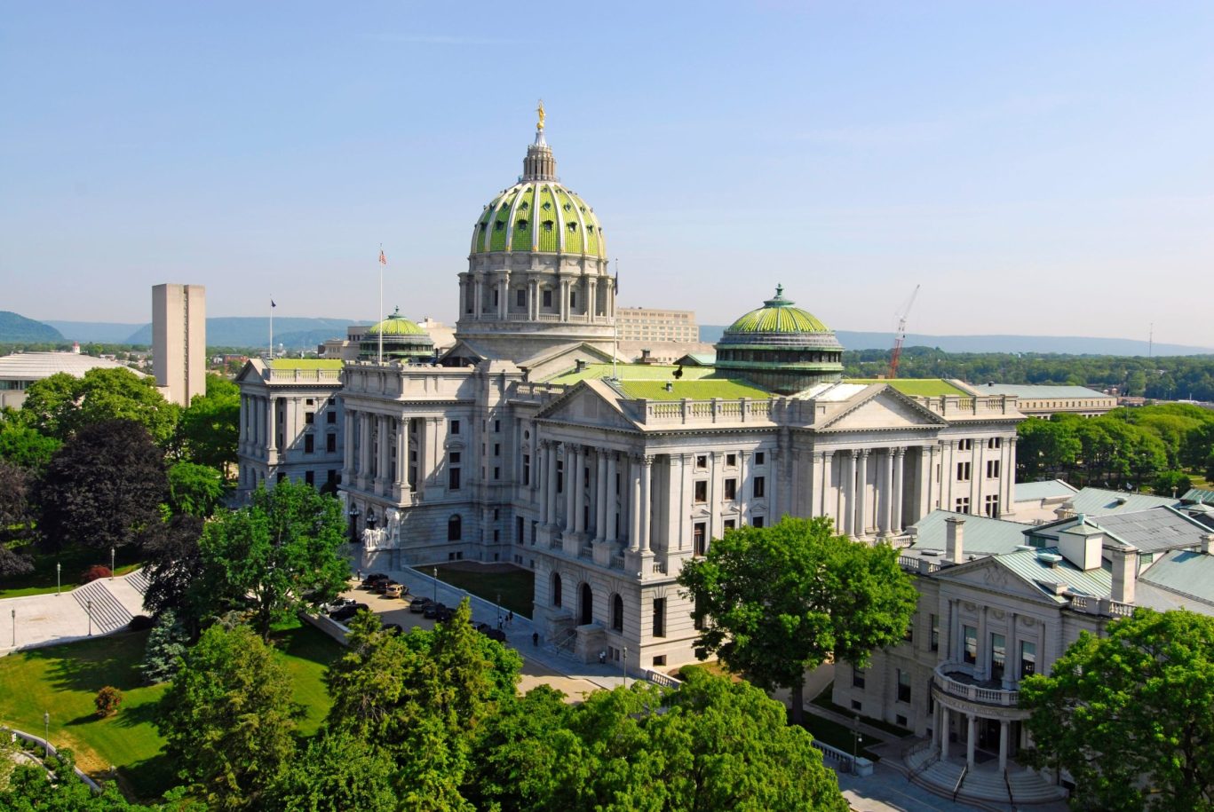 Aerial view of the Pennsylvania State Capitol with green dome and neoclassical architecture surrounded by trees.