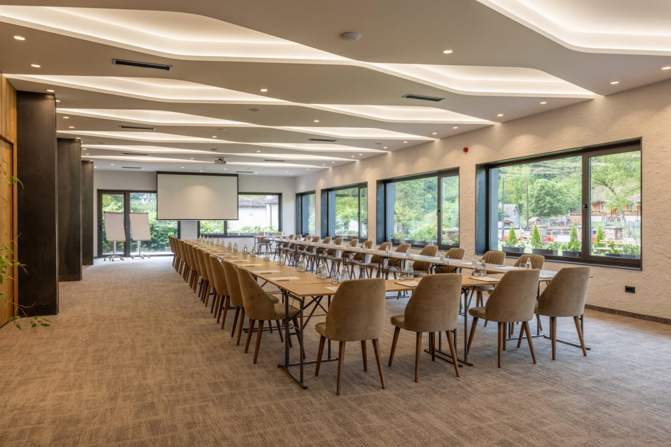 Spacious hotel conference room with large windows, projector screen, and natural lighting.