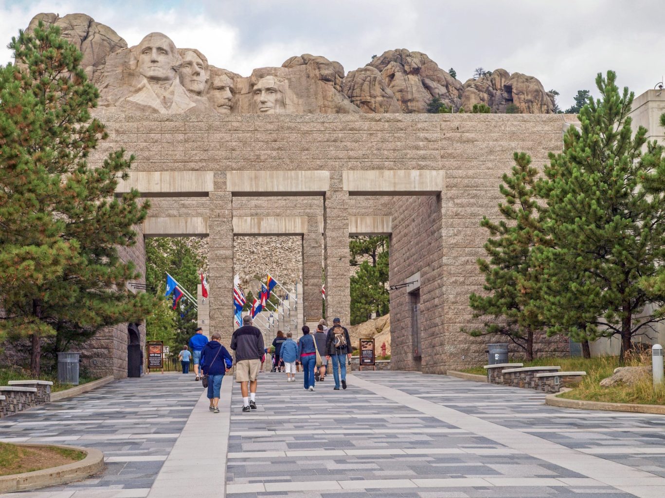 Visitors walking toward Mount Rushmore through the Avenue of Flags entrance surrounded by trees.