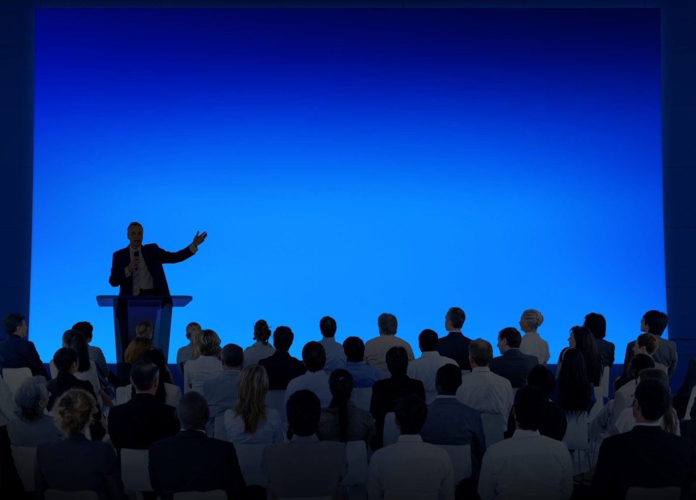 Business conference speaker presenting to a seated audience against a vibrant blue backdrop.