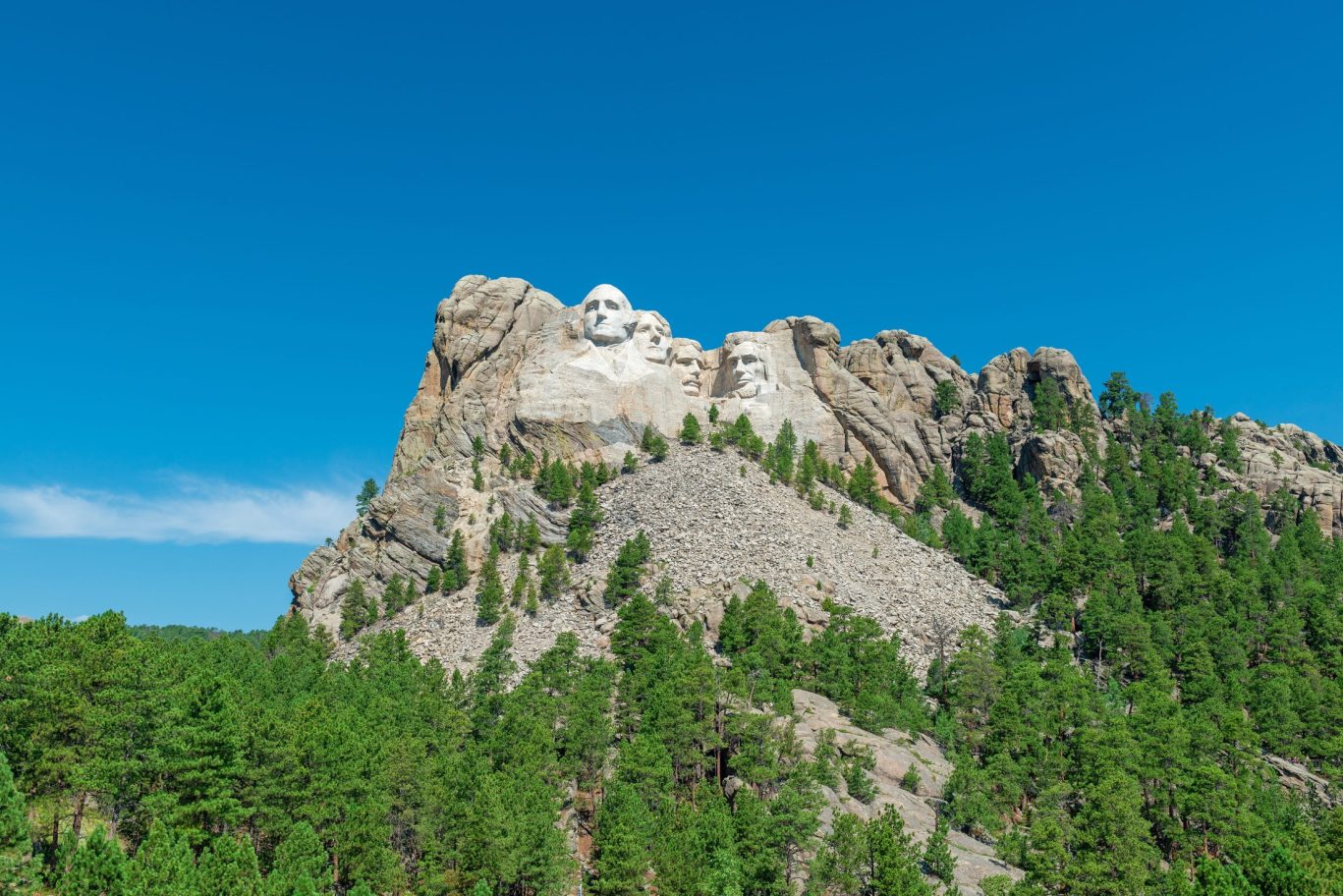 Wide-angle view of Mount Rushmore surrounded by forested hills and bright blue sky.
