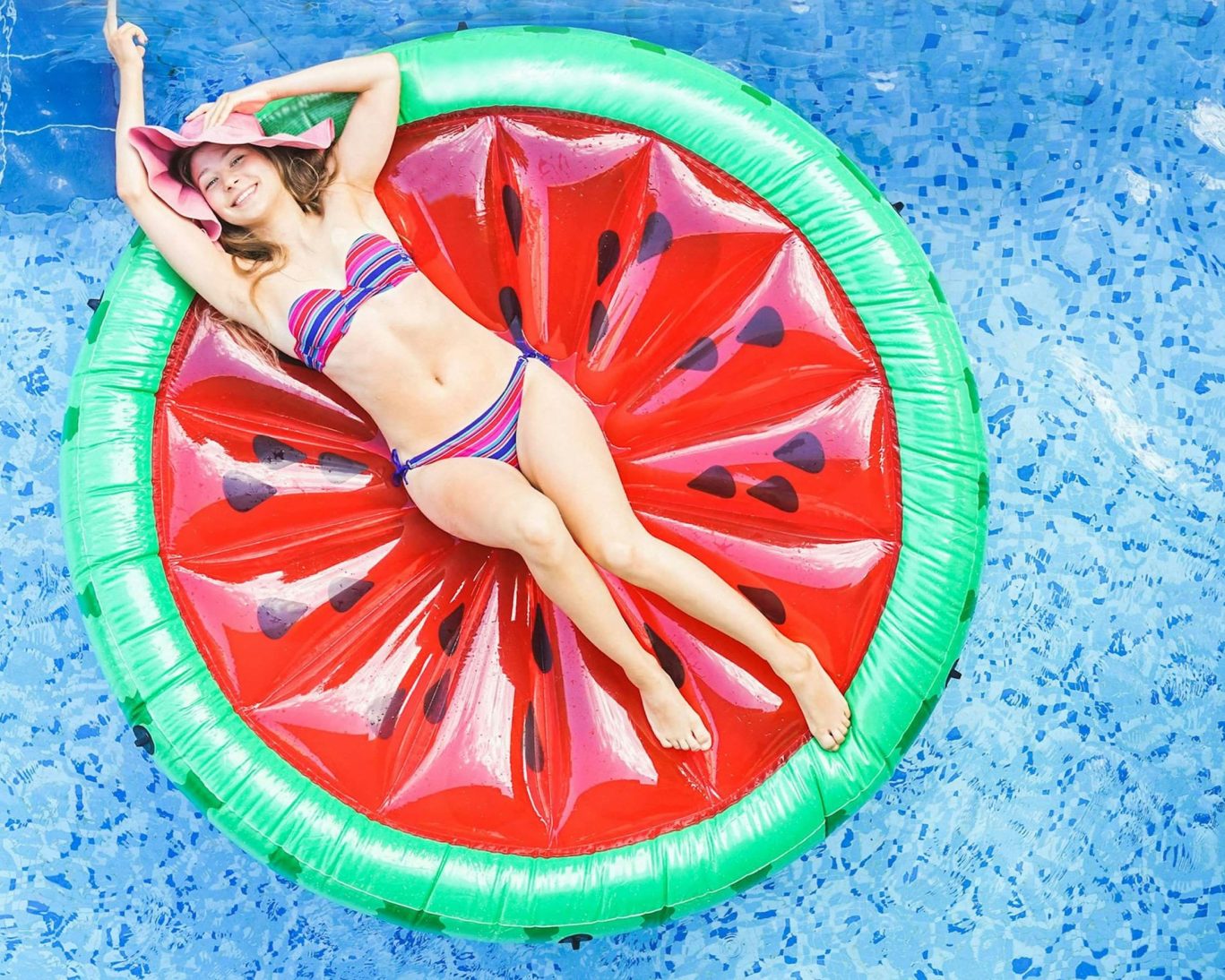 Smiling woman in colorful bikini relaxing on watermelon float in a clear blue swimming pool.