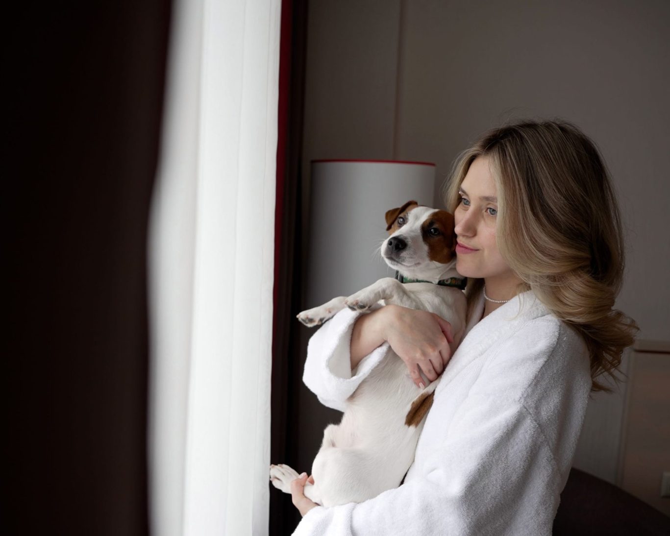 Woman in hotel robe holding pet dog by the window at Magnuson Hotel, highlighting pet-friendly stays.