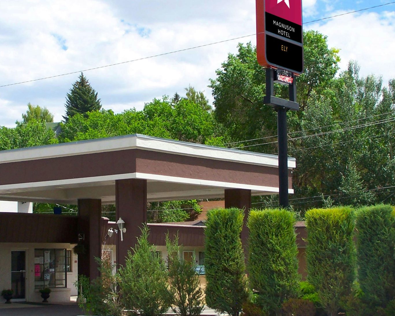Front entrance of Magnuson Hotel Ely surrounded by greenery under a bright blue sky.