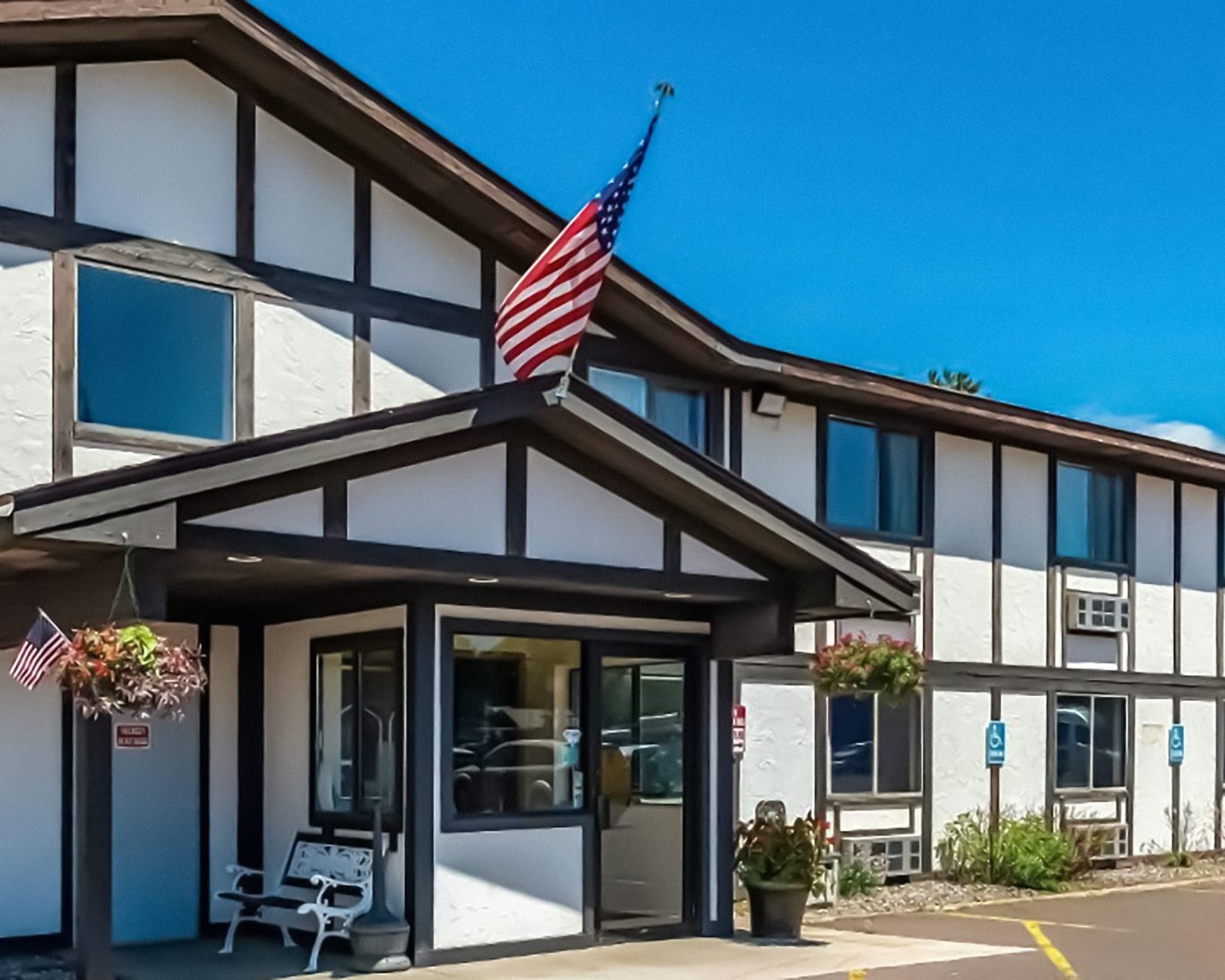 Main entrance of Magnuson Hotel with American flag, accessible parking, and hanging flower baskets.
