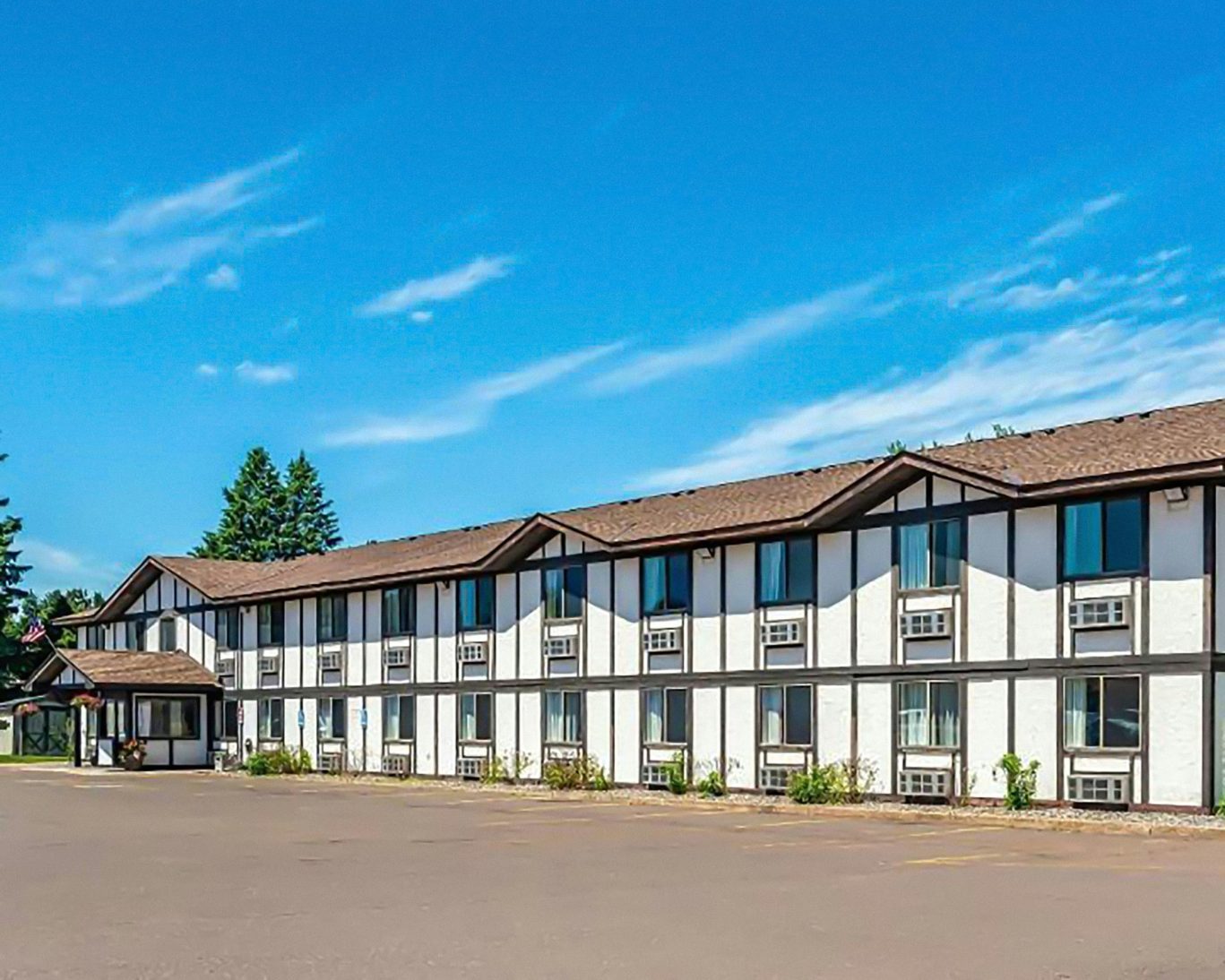 Magnuson Hotel with Tudor-style exterior, individual room A/C units, and large parking lot under blue sky.
