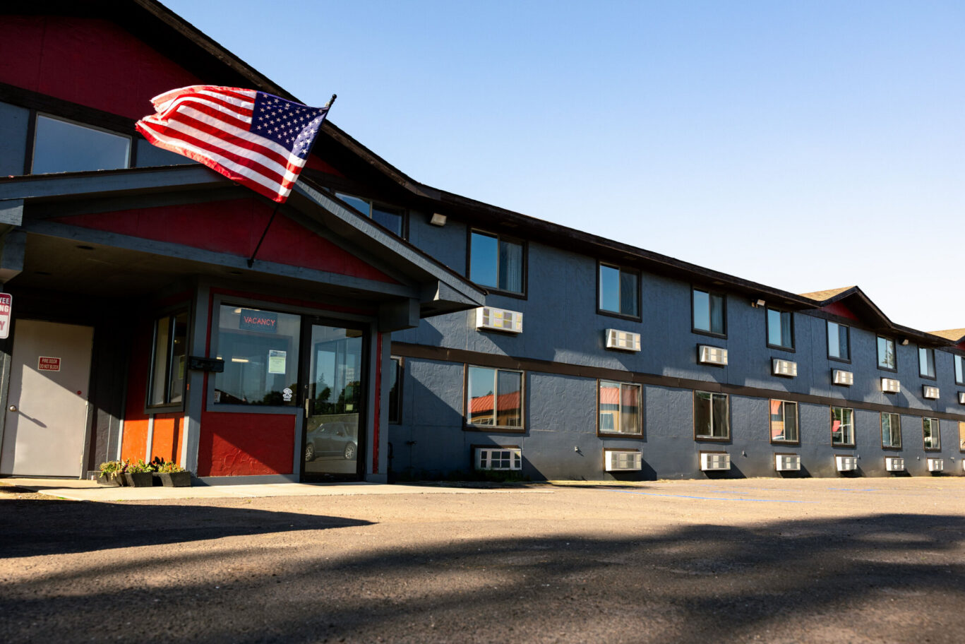 Magnuson Hotel with a Tudor-style exterior, individual room A/C units, and a large parking lot under a blue sky.