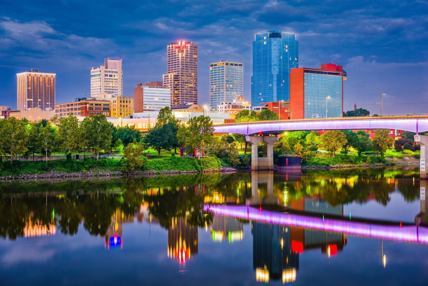 Night view of illuminated Little Rock skyline and River Market District with bridge reflections.