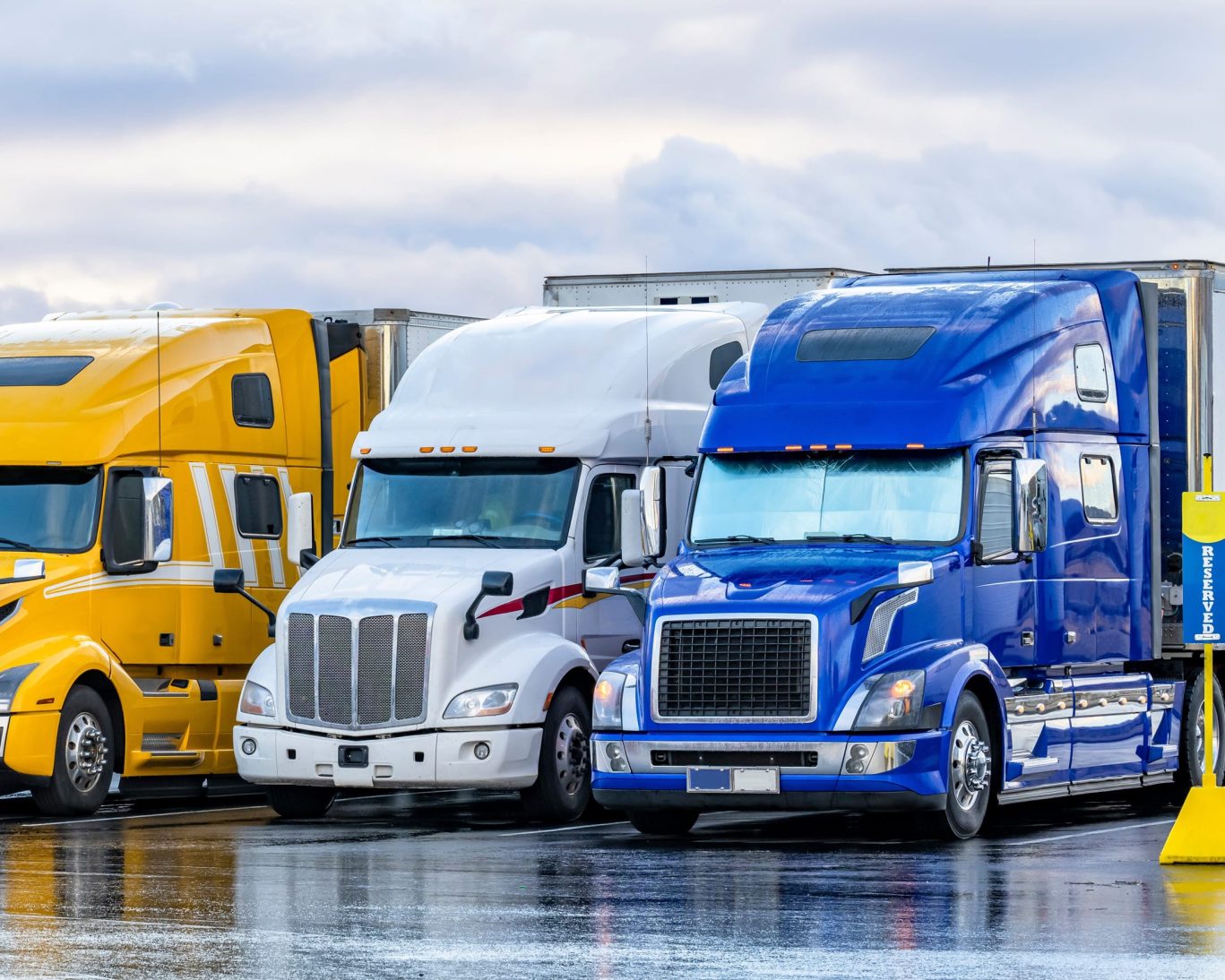 Row of colourful semi-trucks parked in a lot under cloudy skies.