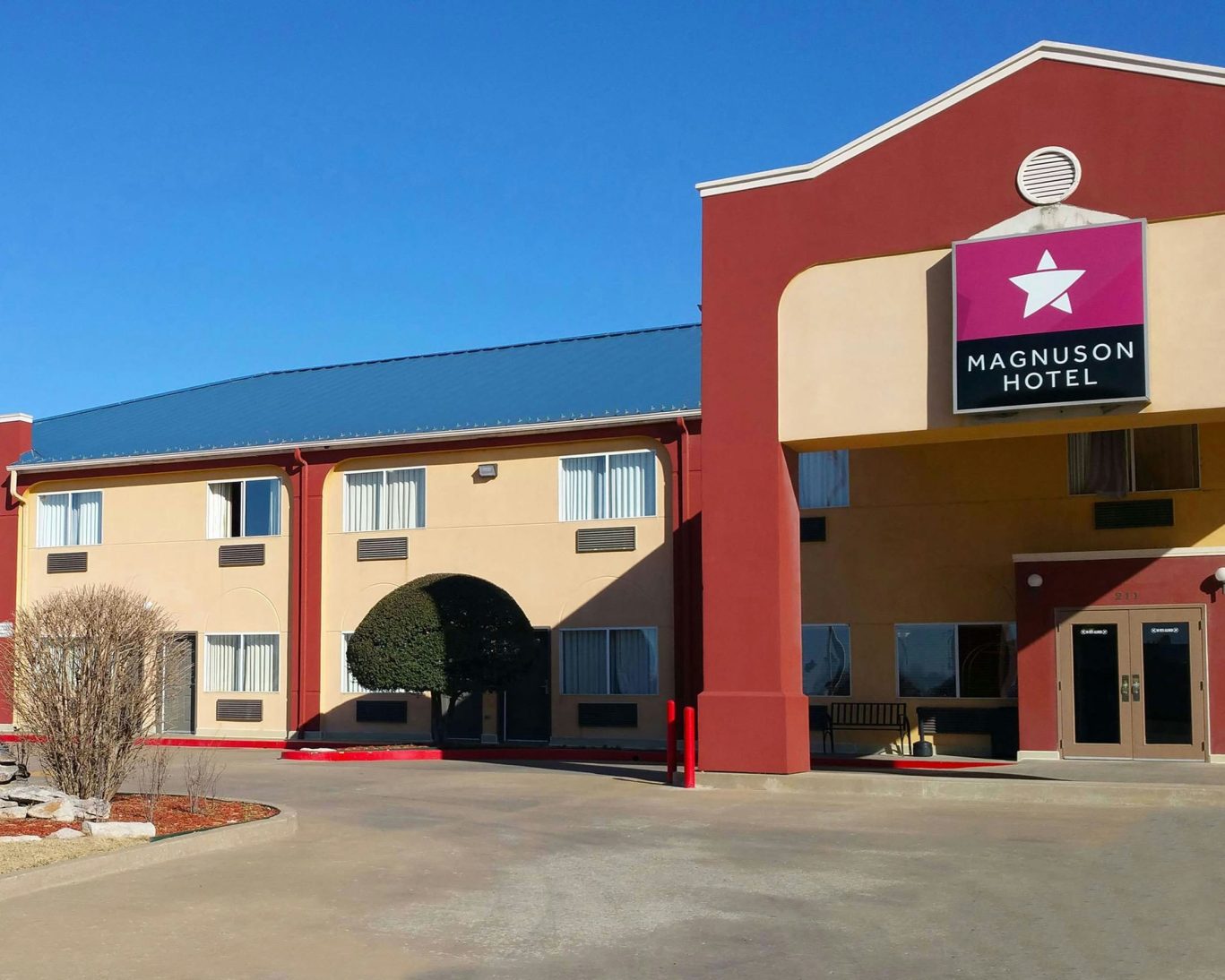 Magnuson Hotel exterior with red and beige facade under clear blue sky.