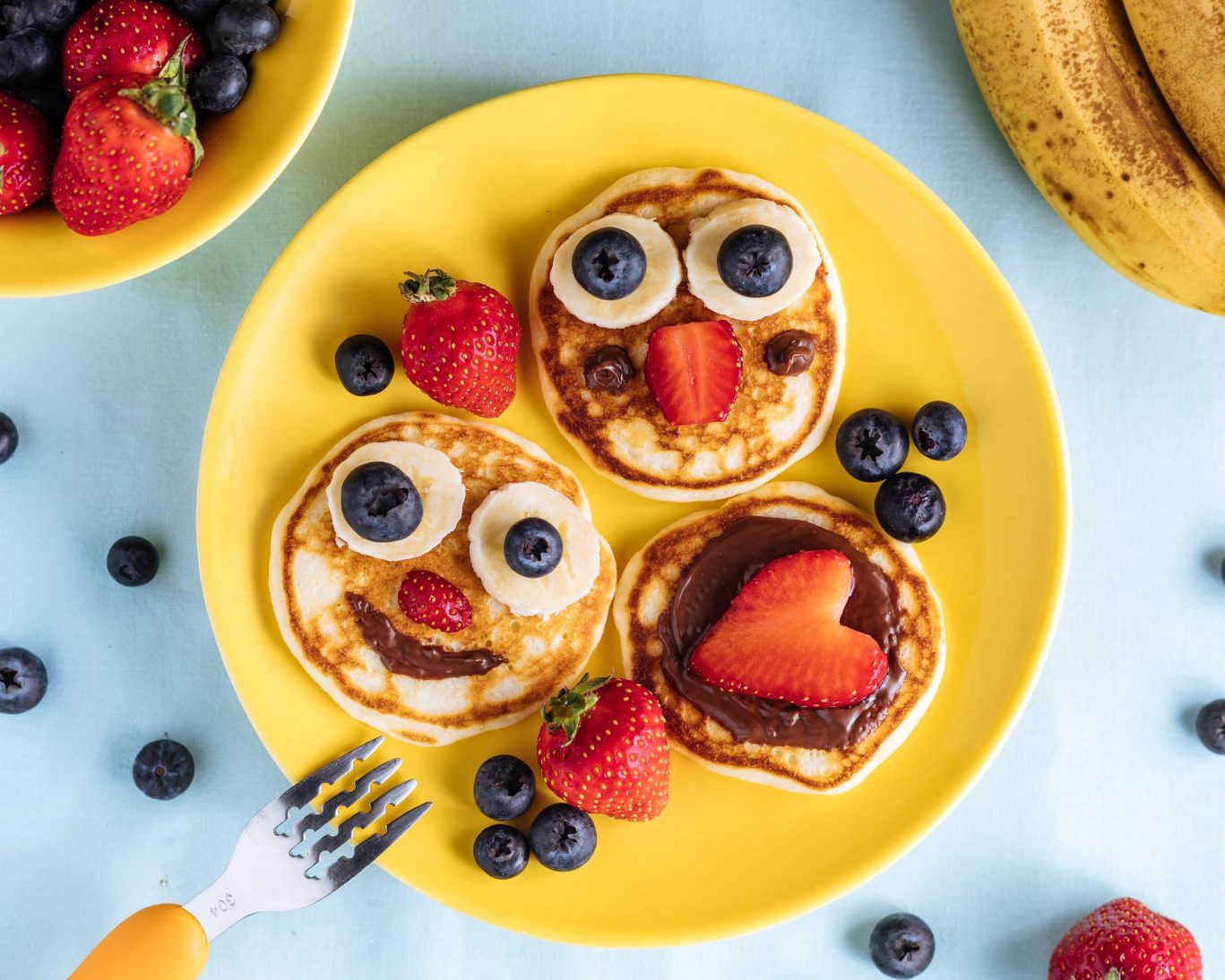 Colorful plate of smiley face pancakes with strawberries, blueberries, and bananas.