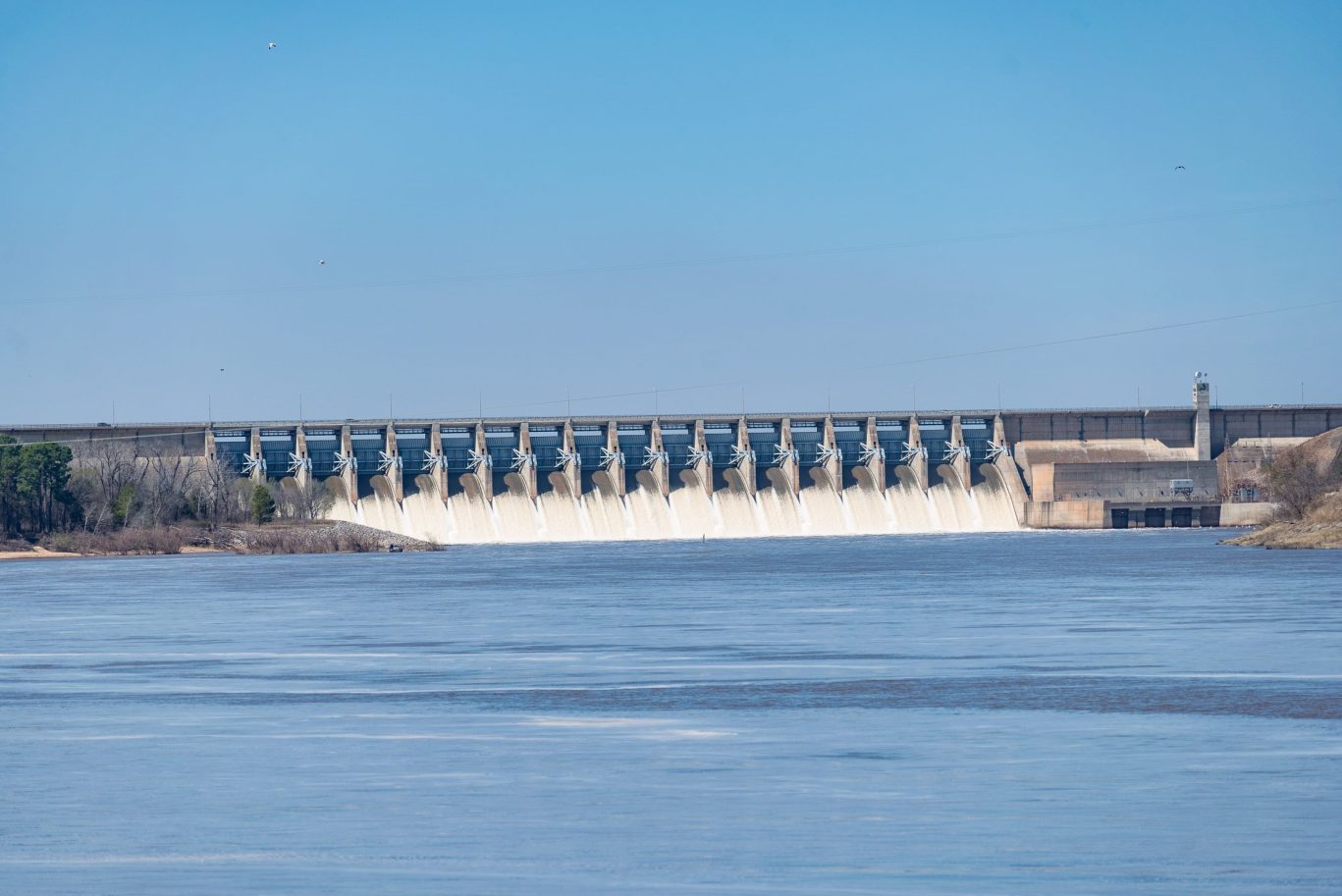 Water flowing from hydroelectric dam with blue river and sky in background.