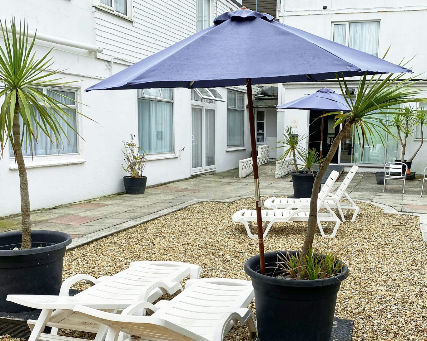 Sunny hotel courtyard with blue umbrellas, white sun loungers, and potted palm trees.