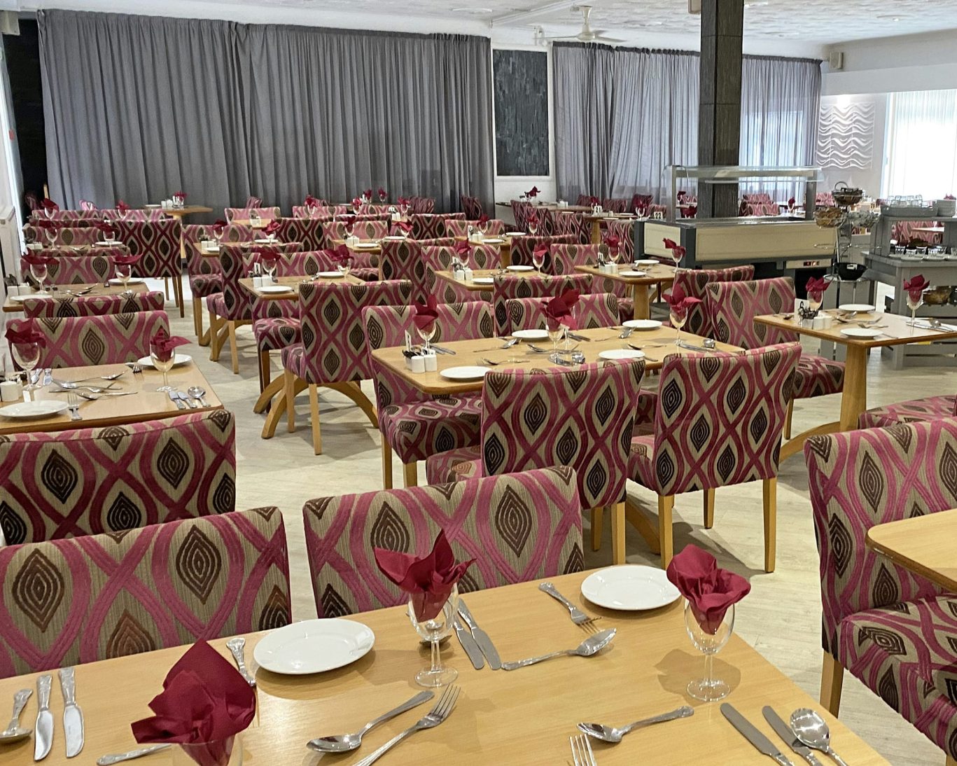 Spacious hotel dining room with red patterned chairs, wooden tables, and formal place settings.