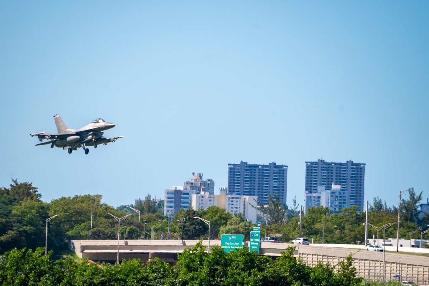 Military fighter jet flying over highway near San Juan, Puerto Rico with city skyline and traffic signs in view.