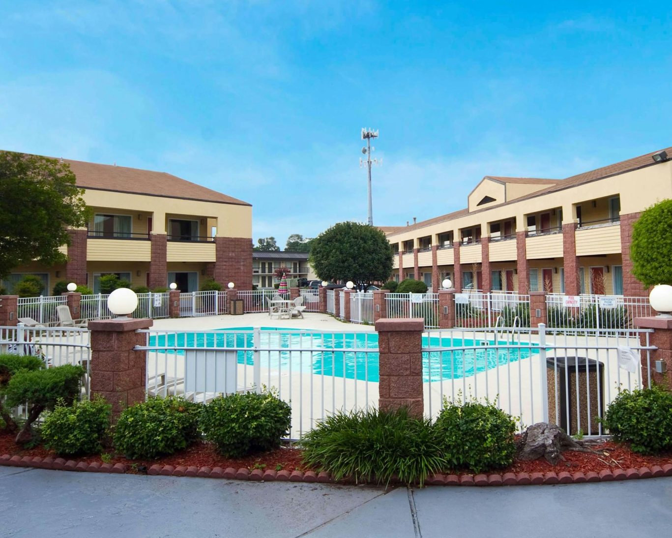 Fenced outdoor hotel pool surrounded by two-story guest rooms and landscaping.
