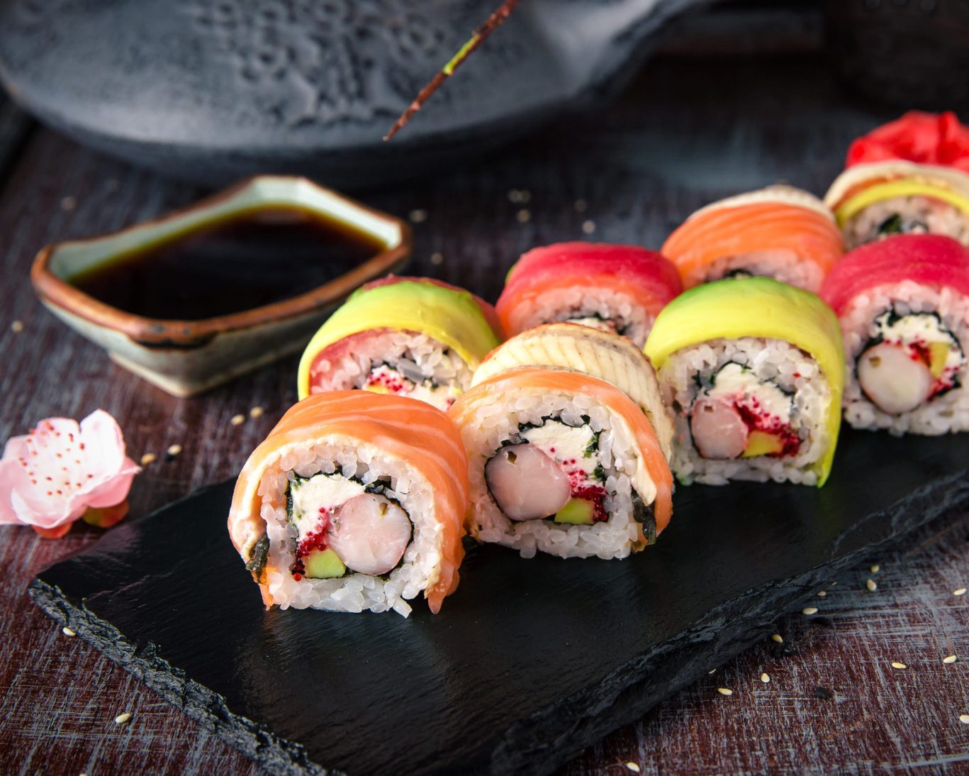 Colorful assorted rainbow sushi rolls on a black slate platter with soy sauce dip.