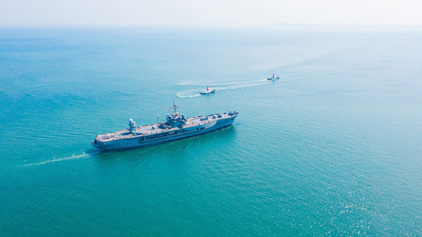 Large military naval ship cruising through calm ocean waters with escort boats.