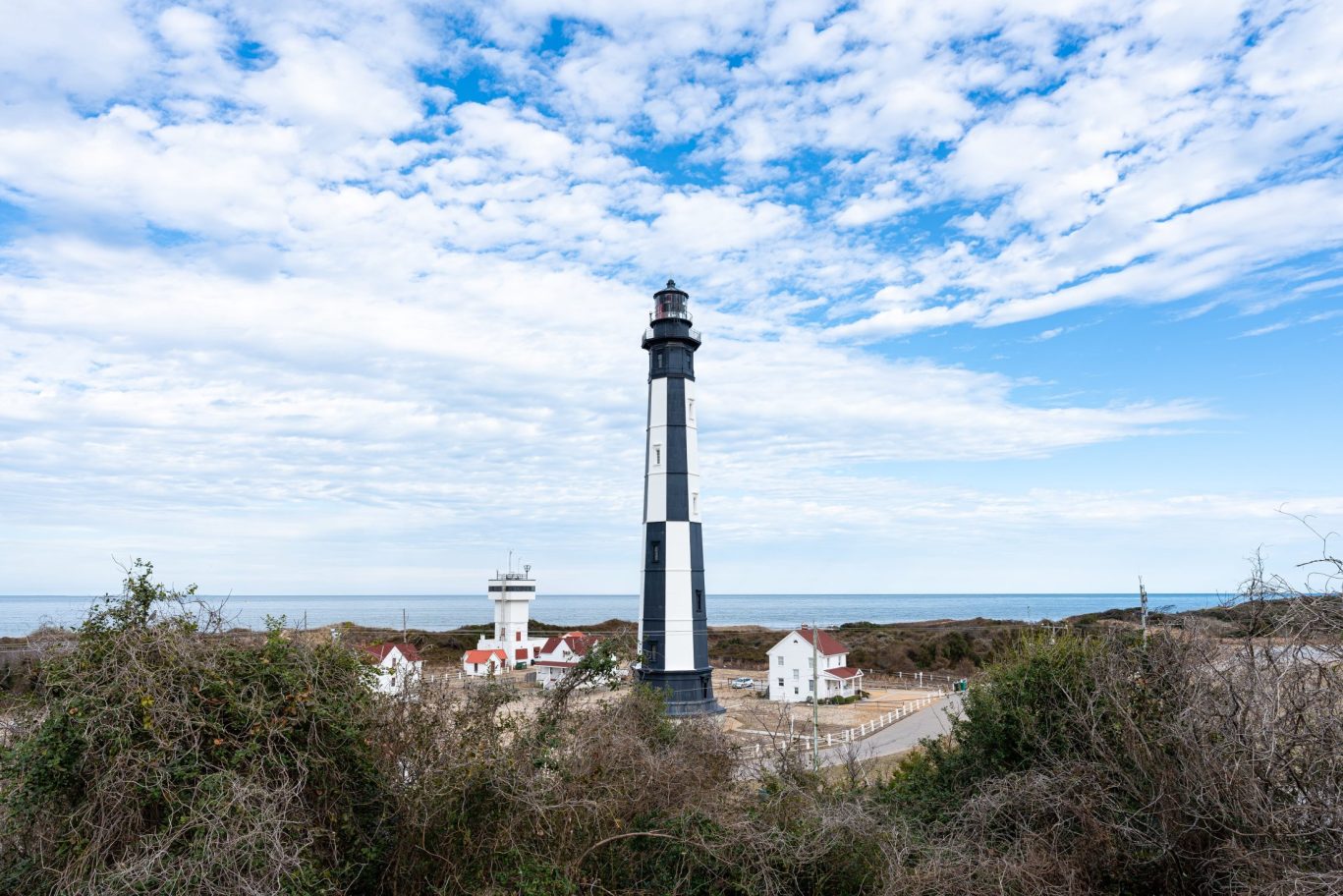 Tall black and white lighthouse with surrounding buildings by the sea under blue sky.