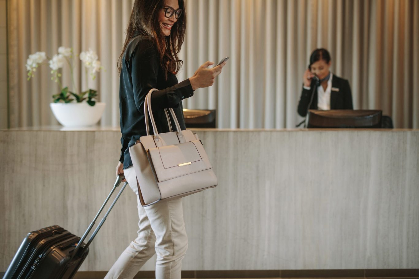 Businesswoman checking into modern hotel lobby with suitcase and smartphone.