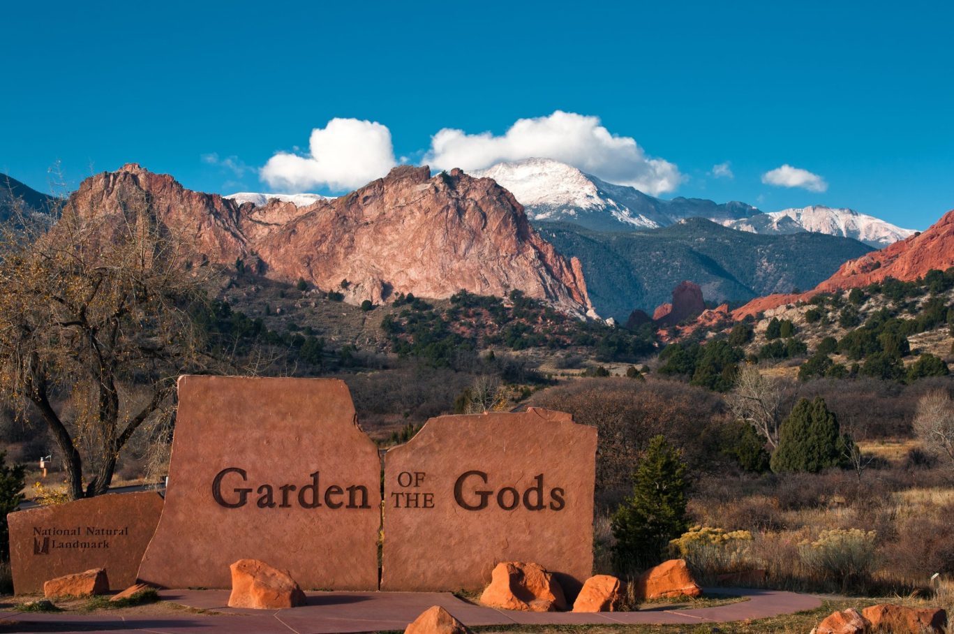 Entrance sign to Garden of the Gods park with red rock formations and mountain view in background.
