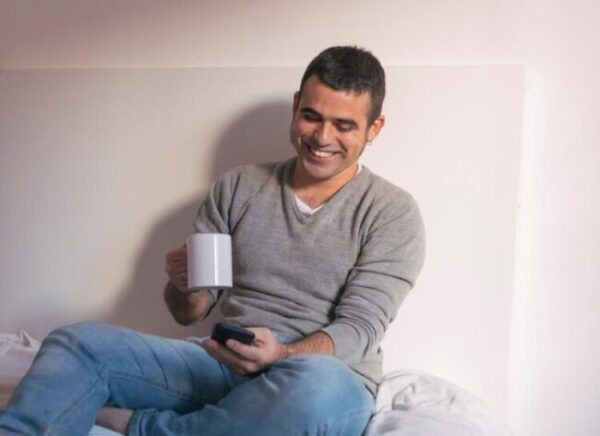 A man sitting cross-legged on a bed, smiling while looking at his smartphone and holding a white mug.