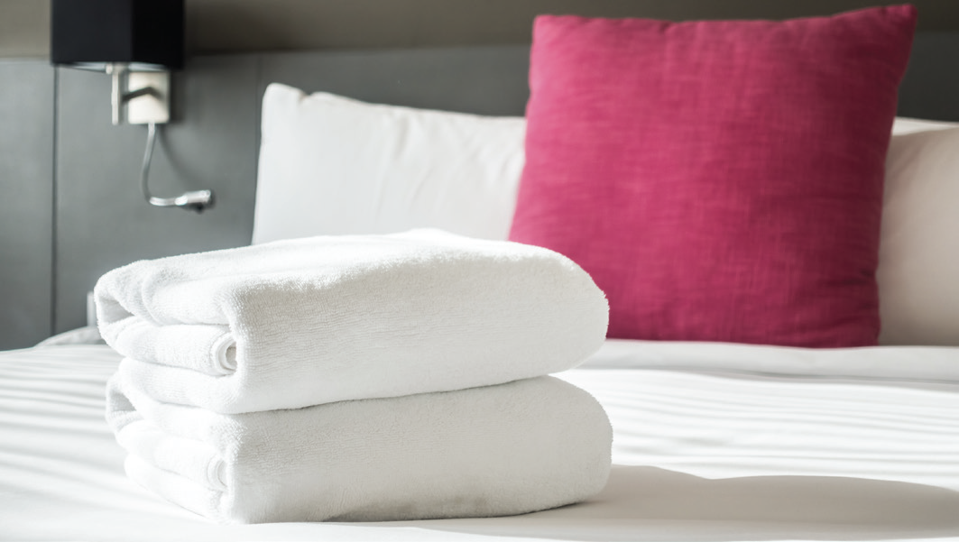 Fresh white towels stacked neatly on a hotel bed with a bright pink / red cushion in the background.