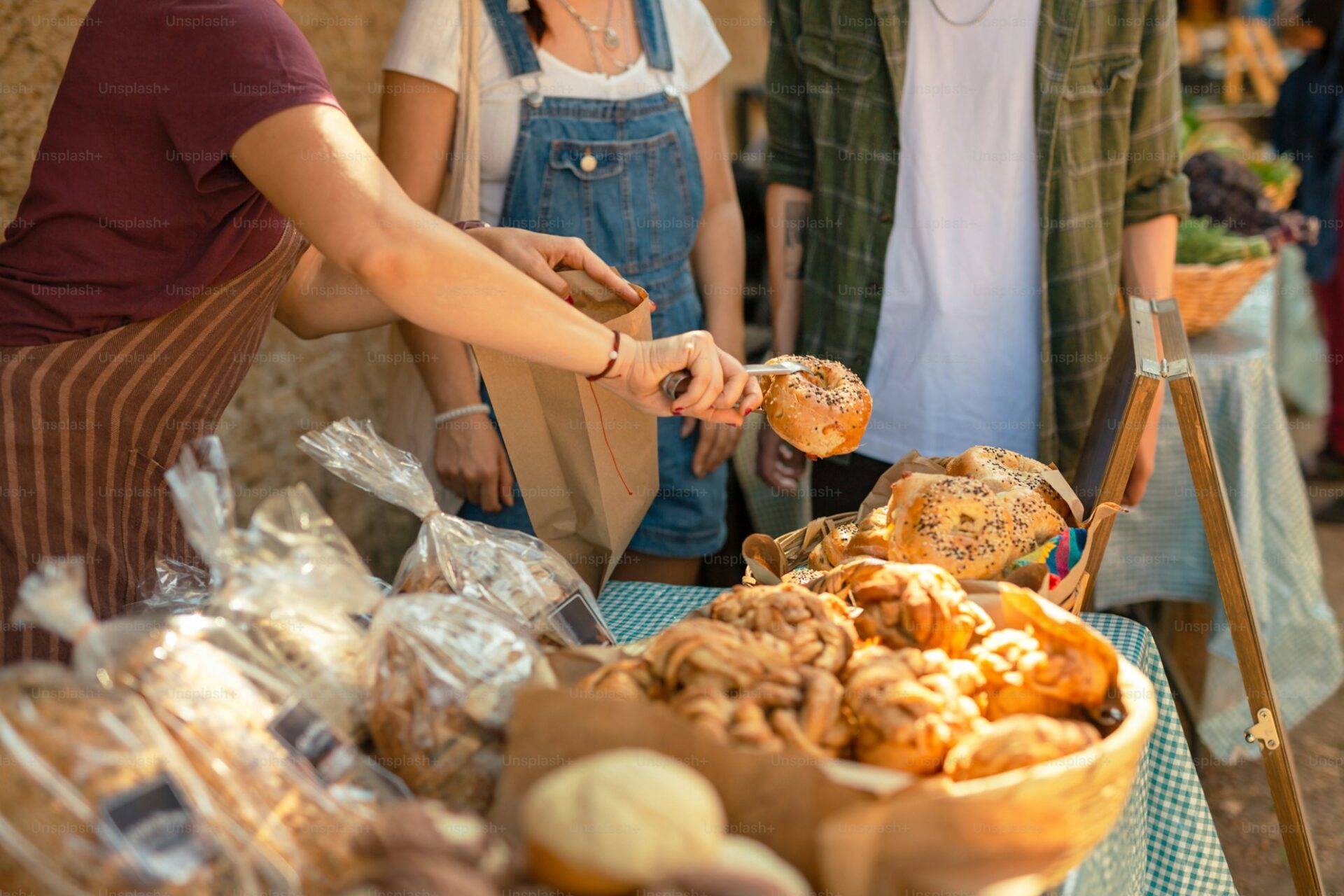 Person placing a freshly baked bagel into a paper bag at an outdoor market stall, with various breads and pastries on display.