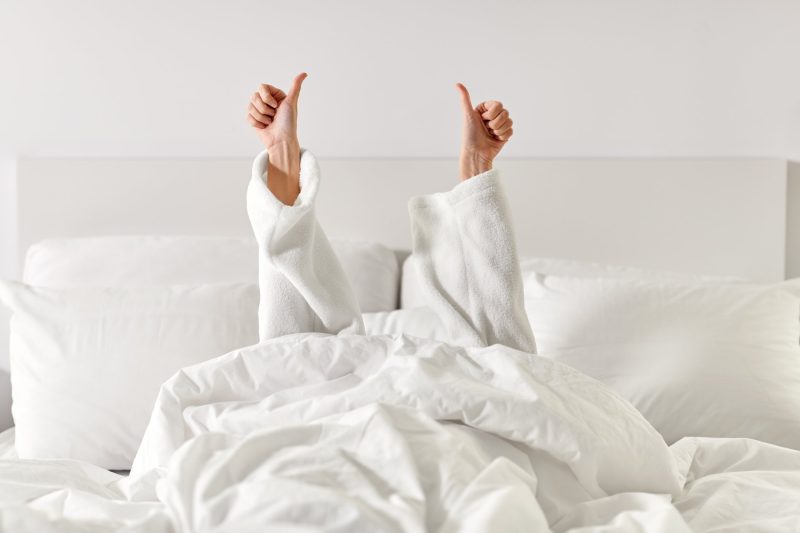 Person in white bathrobe giving two thumbs up while relaxing in a cozy hotel bed with fluffy white linens.