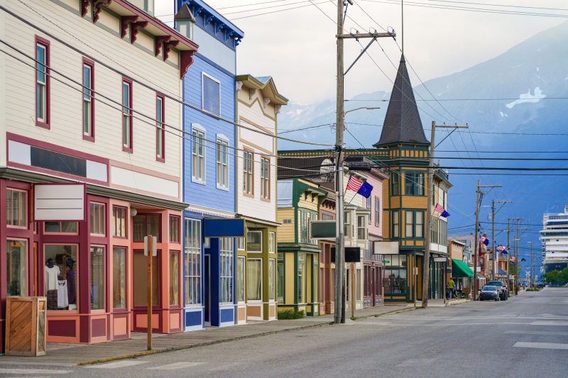 Colorful historic storefronts in downtown Skagway, Alaska with American flags and cruise ship docked in the background.
