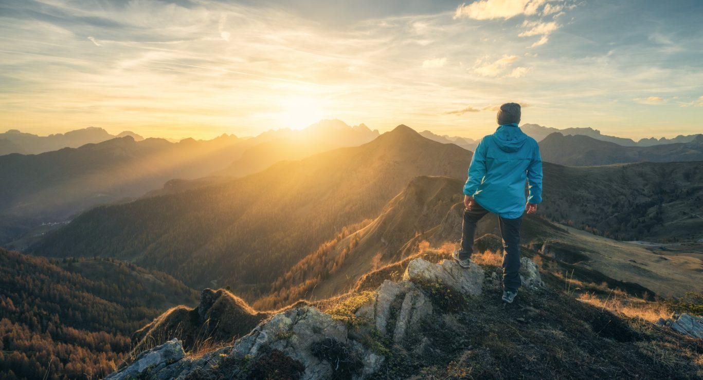 Man standing on a rocky hill overlooking a misty mountain valley at sunset with colorful autumn trees.