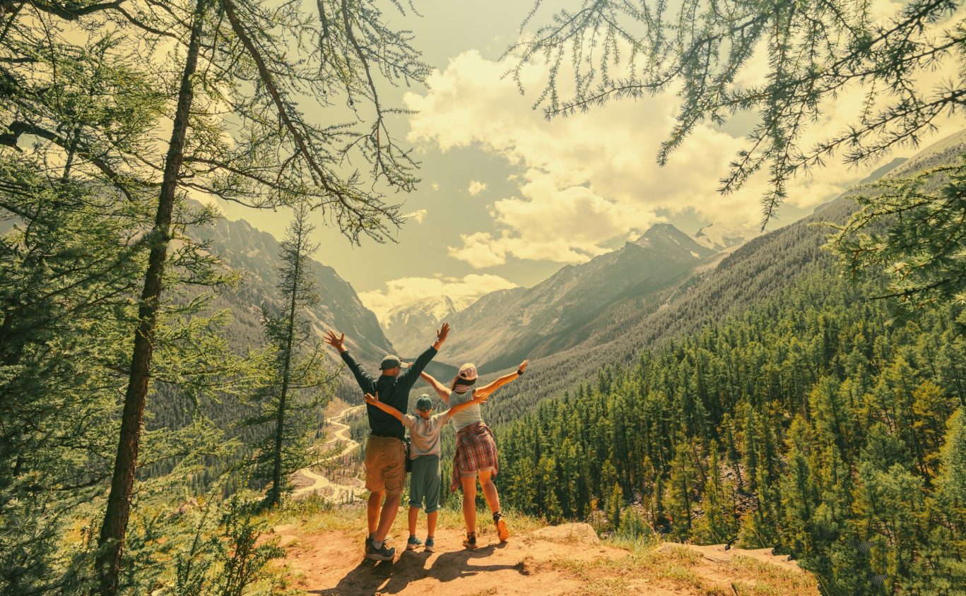 Family of hikers celebrating at a mountain viewpoint surrounded by forest and peaks.