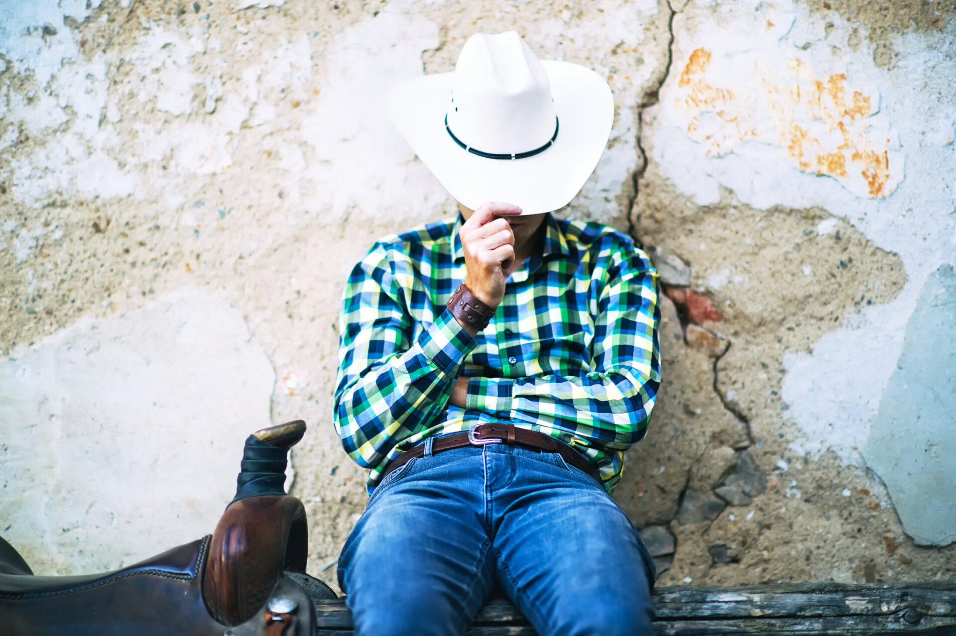 Cowboy in plaid shirt and white hat sitting next to saddle against rustic Texas wall